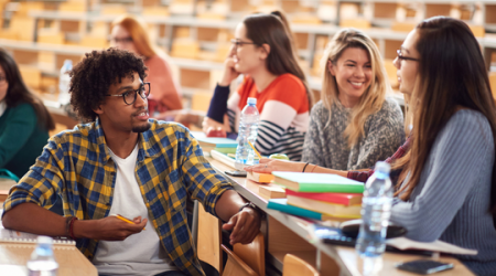 Students seated in rows in a university lecture hall smiling and engaged in relaxed conversation.