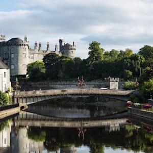 a photo of Kilkenny castle beside river, trees forming a backdrop to stone bridge