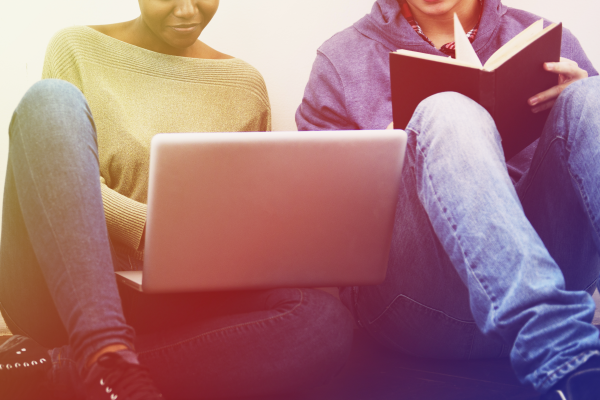 Two college students, one female, the other male, are sitting against a wall; in her lap she is working on a laptop; the male student is reading from a book. Their faces are mostly out of frame.