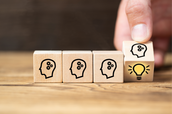Lined up are three small wooden cubes with the engraved outline of a head with cog wheels; a hand is turning a fourth cube at the end of the sequence to reveal a yellow lightbulb, symbolising an idea.