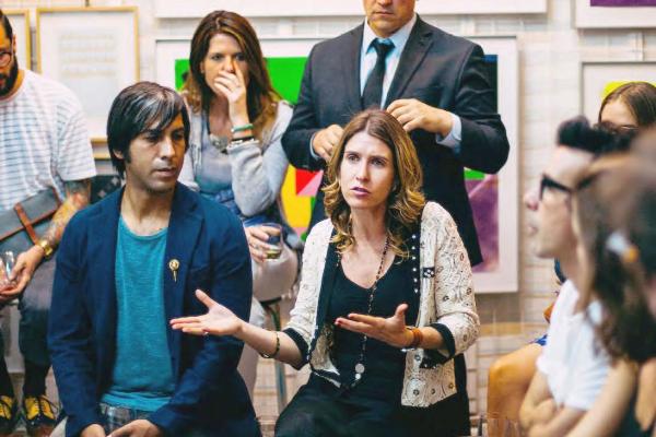 Mature woman seated in art gallery gestures and contributes to gathering of well-attired men and women surrounding her