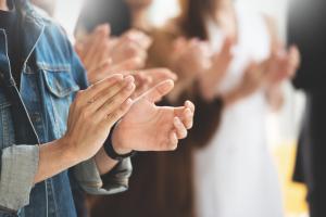 Stock image of pair of hands clapping In foreground; out of focus in mid-distance, other people also clapping indicating a celebration or applause for an achievement
