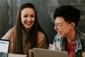 Image of two students laughing together, laptop and notepad on the desk in front of them