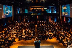 From a rear aspect, a single individual stands in the round addressing a full auditorium