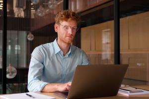 Young professional man, focused, wearing glasses working on laptop, and studying using computer for online seminar webinar at library.