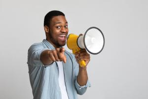 Positive african american guy shouting in megaphone and pointing at camera over grey background