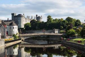 a photo of Kilkenny castle beside river, trees forming a backdrop to stone bridge