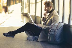 alt="Young man sitting indoors at an airport using laptop."